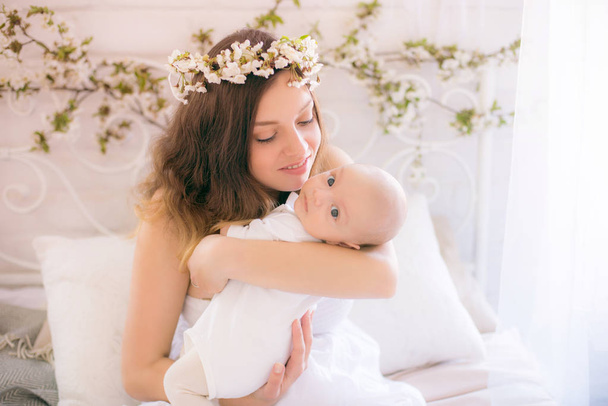 Young cute mom in a wreath of cherry blossoms in a white dress holding a baby in her arms in a spring bedroom with blossoming cherry branches - Foto, Bild