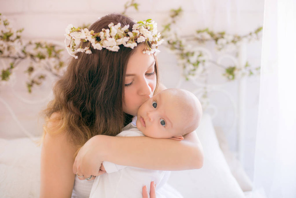 Young cute mom in a wreath of cherry blossoms in a white dress holding a baby in her arms in a spring bedroom with blossoming cherry branches - Foto, imagen