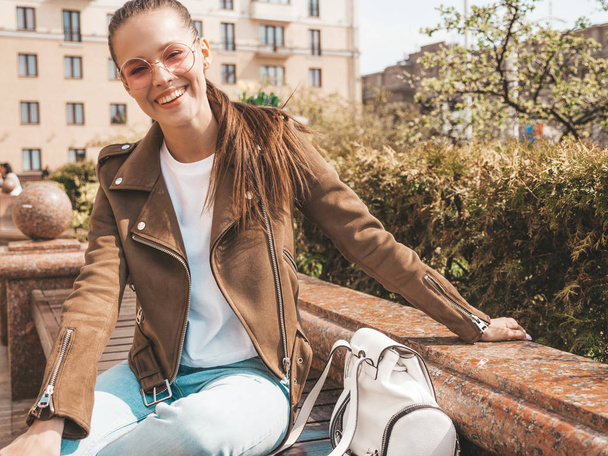 Portrait of beautiful smiling brunette model dressed in summer hipster jacket and jeans clothes. Trendy girl sitting on the bench in the street background. Funny and positive woman in round sunglasses - Zdjęcie, obraz