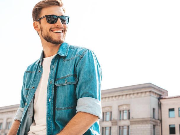Portrait of handsome smiling stylish hipster lumbersexual businessman model. Man dressed in jeans jacket clothes. Fashion male posing on the street background in sunglasses - 写真・画像