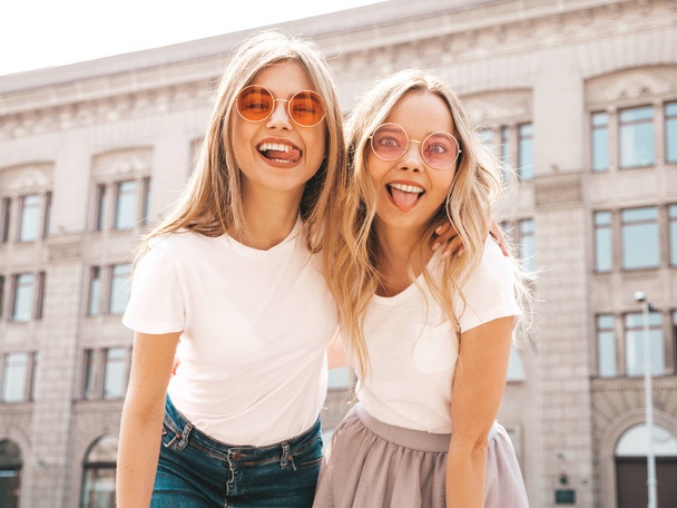 Porträt zweier junger hübscher blonder lächelnder Hipstermädchen in trendiger sommerweißer T-Shirt-Kleidung. sexy unbeschwerte Frauen posieren auf der Straße Hintergrund. Positive Models, die ihre Zunge zeigen, mit Sonnenbrille - Foto, Bild