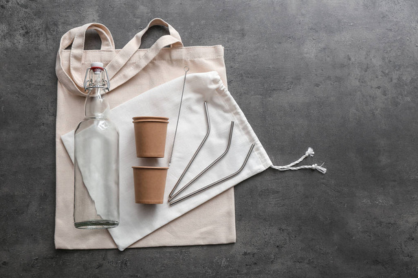 Стеклянная бутылка с пакетами, чашками и соломинками на сером фоне. Концепция нулевых отходов
 - Фото, изображение