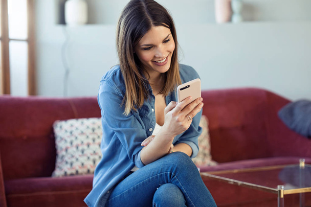 Pretty young woman using her mobile phone while sitting on sofa at home. - 写真・画像