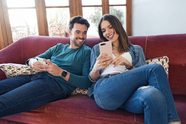 Lovely young couple using they mobile phone while sitting on sofa at home. - Fotó, kép