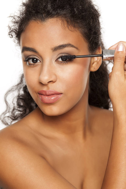 portrait of a young dark-skinned woman applying mascara on a white background - Photo, Image