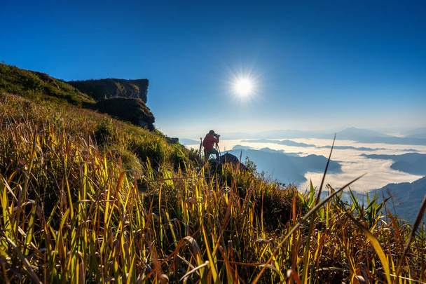 Фотограф держит камеру в руке и стоит на вершине скалы в природе. Phu chi fa mountains in Chiang rai, Thai
 - Фото, изображение