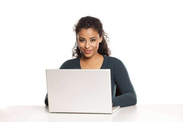 Gülümseyen ve pozitif genç afrikalı-amerikalı kadın güzel yüzü ile dizüstü bilgisayar kullanarak, beyaz arka plan üzerinde masada proje çalışma - Fotoğraf, Görsel