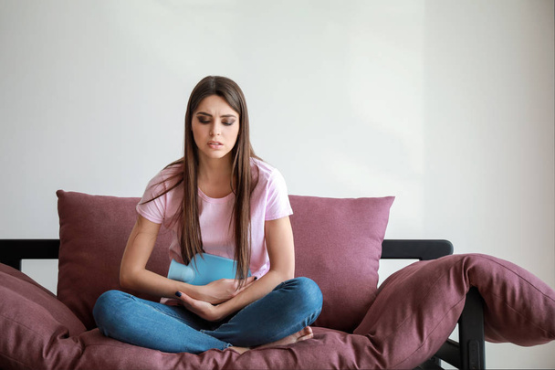 junge Frau leidet unter Bauchschmerzen, während sie auf dem Sofa vor weißem Hintergrund sitzt - Foto, Bild