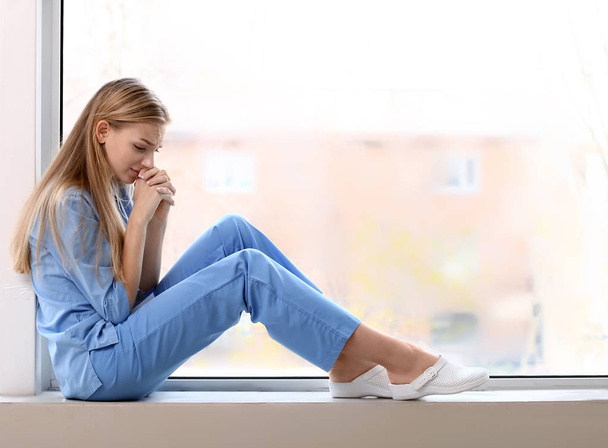 Depressed female medical assistant sitting on window sill in clinic - Zdjęcie, obraz