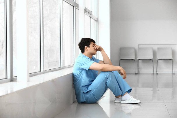 Depressed male medical assistant sitting near window in clinic - Foto, Imagen