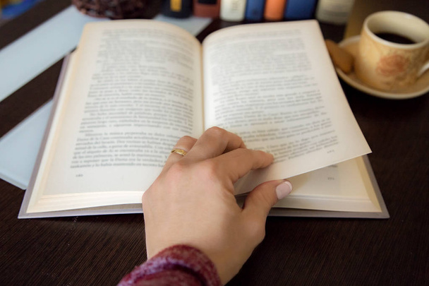 kadın el arka planda daha fazla kitap ve kahve fincan ile koyu ahşap kitap bir sayfa dönüm detay - Fotoğraf, Görsel