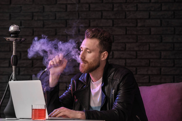 junger Mann raucht Wasserpfeife, während er im Café am Laptop arbeitet - Foto, Bild