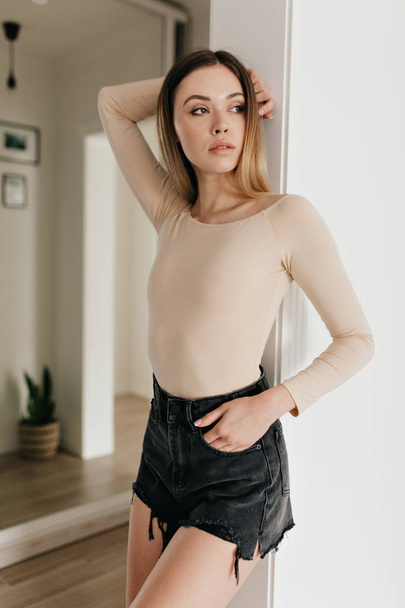 Increíble mujer joven elegante con cuerpo beige y pantalones cortos posando durante la sesión de fotos en el apartamento moderno
  - Foto, imagen