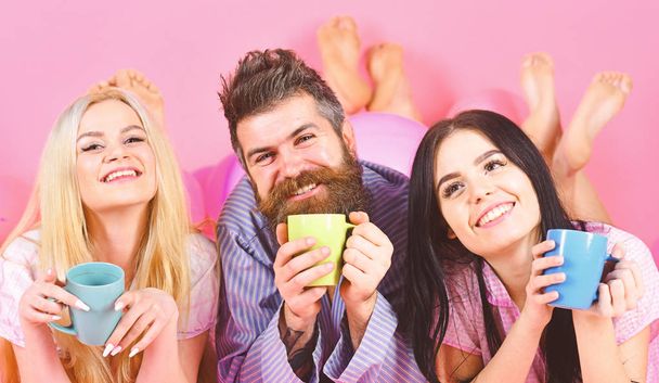 Lovers drinking coffee in bed. Man and women, friends on smiling faces lay, pink background. Man and women in domestic clothes, pajamas. Threesome relax in morning with coffee. Lovers in bed concept - Photo, image