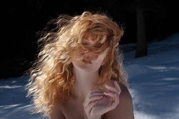 Young sexy blonde naked woman in the winter, after the sauna, wellness, covered in snow, freezes and looks out with one eye - 写真・画像