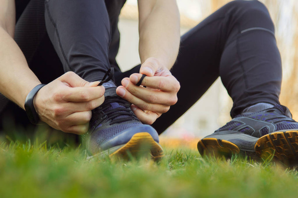 A young man in black clothes is tying the laces on the sneakers close up. fitness athlete sitting on the sports field on the grass. warm up body preparation for the training - Foto, Imagen
