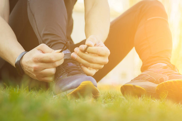 A young man in black clothes is tying the laces on the sneakers close up. fitness athlete sitting on the sports field on the grass. warm up body preparation for the training - Фото, изображение