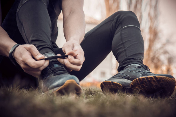 A young man in black clothes is tying the laces on the sneakers close up. fitness athlete sitting on the sports field on the grass. warm up body preparation for the training - 写真・画像
