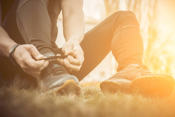 A young man in black clothes is tying the laces on the sneakers close up. fitness athlete sitting on the sports field on the grass. warm up body preparation for the training - Photo, Image