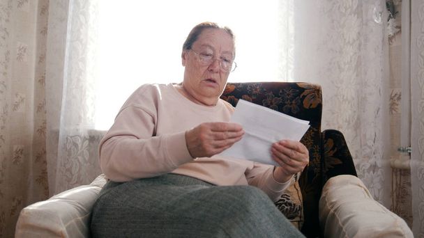 Stara zdumiona Kobieta w okularach siedzi na fotelu i trzyma rachunki za narzędzia - Zdjęcie, obraz