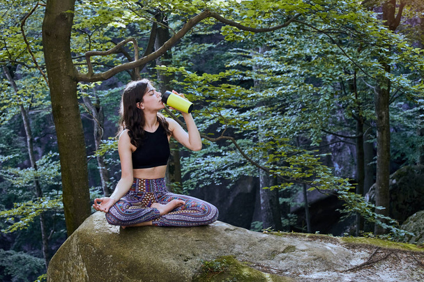 Healthy fitness girl sitting in lotus pose and drinking water from green bottle in forest - Foto, immagini
