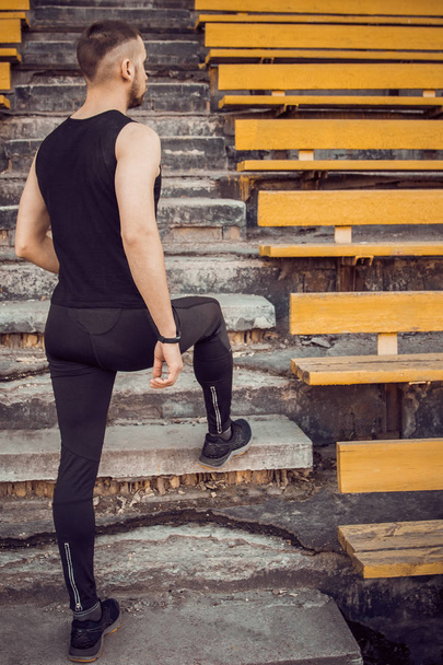 A slender young man in black sportswear stands in front of the stairs stadium stands. ready for action, step to victory, challenge. vertical - Photo, Image