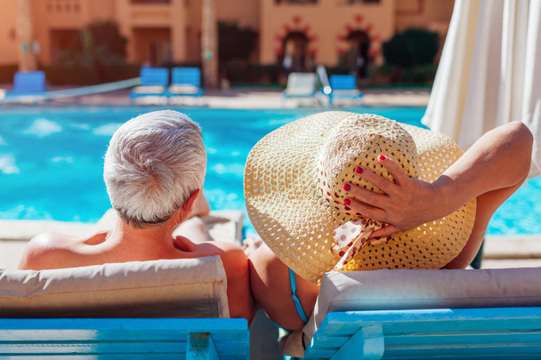Senior couple relaxing by swimming pool lying on chaise-longues. People enjoying summer vacation. - Photo, Image