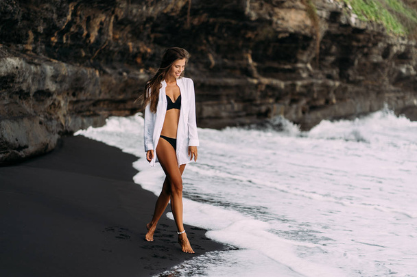 Young sexy girl in white shirt and swimsuit resting on black sand beach. - Φωτογραφία, εικόνα