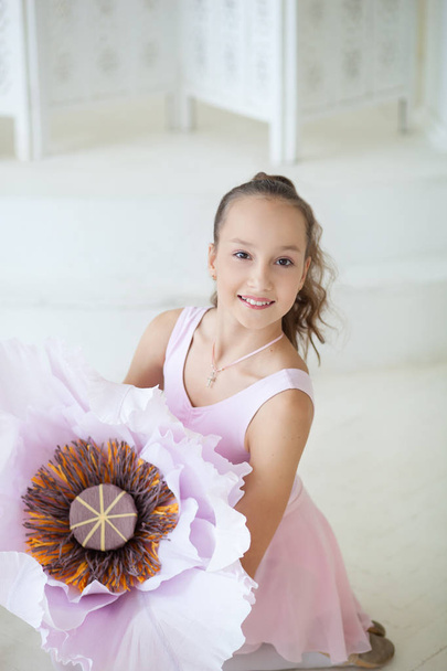 Bir bale kostümü sevimli küçük balerin büyük bir çiçek ve gülümseyerek katta oturuyor. Dans sınıfındaki kız. Kız bale okuyor. Klasik bale. Büyük çiçekler. Bale stüdyosu - Fotoğraf, Görsel
