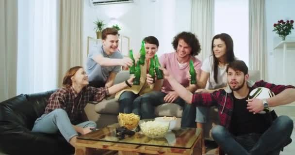 Very enthusiastic friends guys and ladies enjoying the time together in front of the camera watching a football match and supporting their best team drinking beer and cheers with bottles - Materiał filmowy, wideo