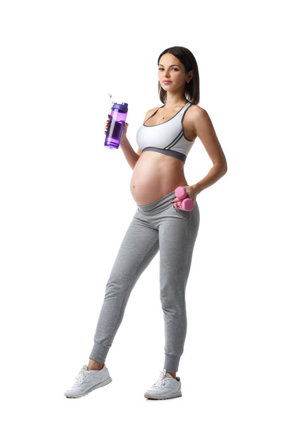 schlanke athletische schwangere Mädchen steht mit einer Flasche Wasser und einer Hantel in der Hand isoliert auf weißem Hintergrund. - Foto, Bild