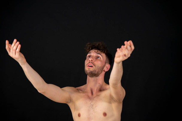 Retrato semidesnudo de un joven bailarín, en movimientos relacionados con el baile con las manos. El joven atractivo con barba en demostraciones de danza moderna sobre un fondo negro
. - Foto, Imagen