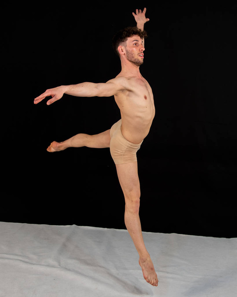 Un danseur masculin portant une coulotte de couleur chair pratique des mouvements de danse aérienne en studio. Le jeune homme musclé attrayant dans les démonstrations conceptuelles de la danse moderne sur un fond noir
. - Photo, image