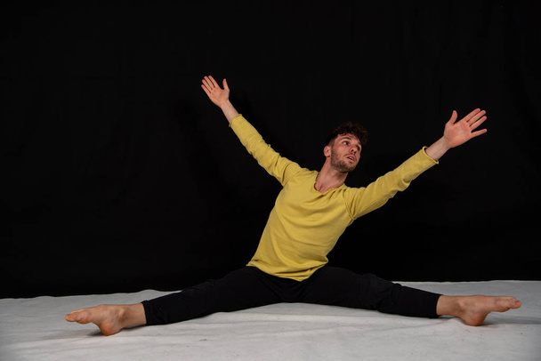 Joven bailarín masculino en movimientos de baile. Figuras con el cuerpo sentado en el suelo sobre un fondo negro. El joven en demostraciones conceptuales de danza moderna
. - Foto, Imagen
