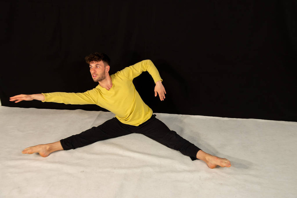 Dançarino masculino com calças pretas e camiseta amarela, praticando no estúdio em fundo preto. O jovem atraente mostra poses de dança moderna
. - Foto, Imagem