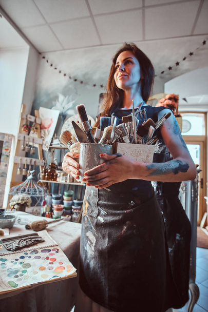 Joven pintora atractiva posa con pinceles en su propio taller acogedor
 - Foto, Imagen