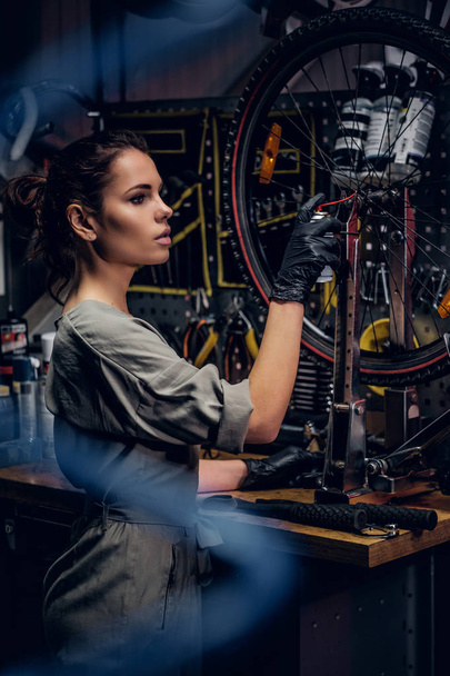 Beautiful woman is repairing bicycle at busy workshop between pneumatic wires. - Photo, Image
