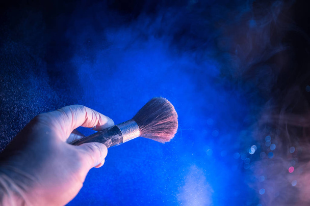 Makijaż w ręku szczotka z kosmetycznych proszku na ciemnym tle z światła i dymu. Proszku splash na ciemny. Selektywny fokus - Zdjęcie, obraz