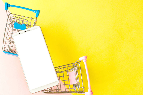 Blank screen white cellphone and shopping carts on pastel pink and yellow background. Minimal style, flatlay - Photo, Image