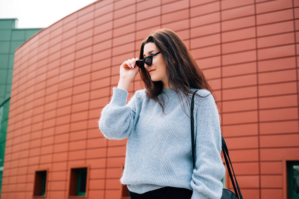 Beautiful young girl with dark hair wearing casual clothes posing near red wall, street style, outdoor portrait, hipster girl in sunglasses. Stylish millennial woman in city street, urban concept. - Φωτογραφία, εικόνα
