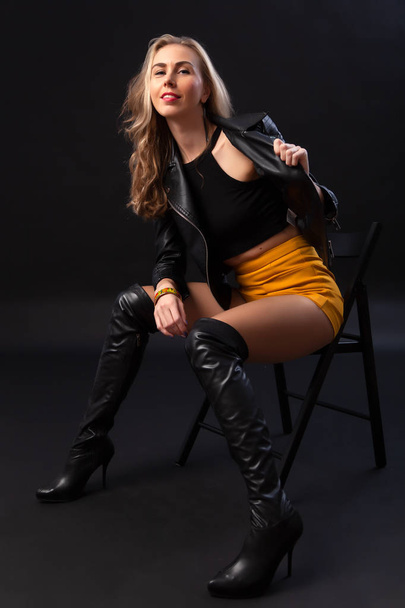 Beautiful woman with healthy body wearing in a black leather jacket, black top, short skirt and boots on black background - Photo, Image