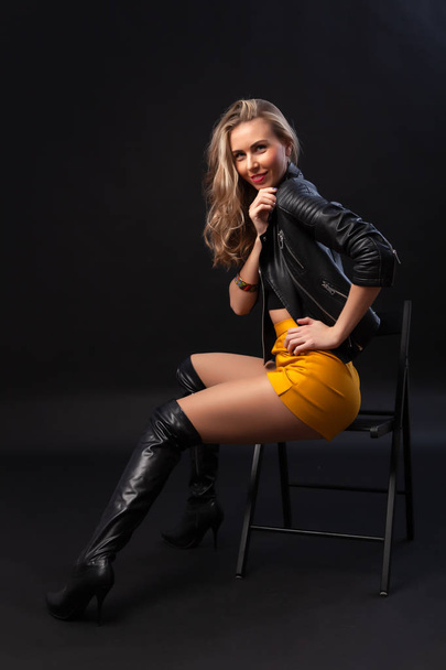 Young beautiful woman wearing in a black leather jacket, black top, short skirt and boots  posing on chair over black background. Studio shot of  blonde woman against black background - Photo, image