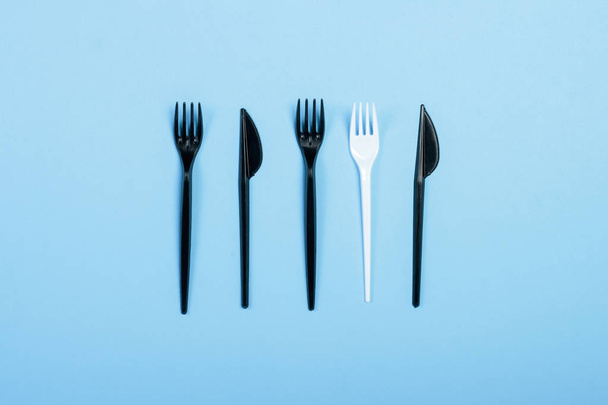 Tenedores y cuchillos de plástico negro y un tenedor de plástico blanco sobre un fondo azul. Concepto plástico, dañino, contaminación ambiental, detener el plástico. Piso tendido, vista superior
. - Foto, imagen
