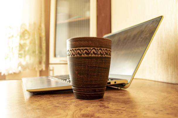 глиняная чашка с вареным кофе на кухонном столе возле портативного компьютера рано утром, работа в неформальной обстановке
 - Фото, изображение