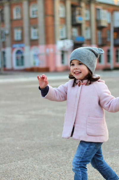 mooi klein meisje in een hoed, jas en jeans springt, vliegt en heeft plezier op de achtergrond van het stadsbeeld in het licht van de zonsondergang. - Foto, afbeelding