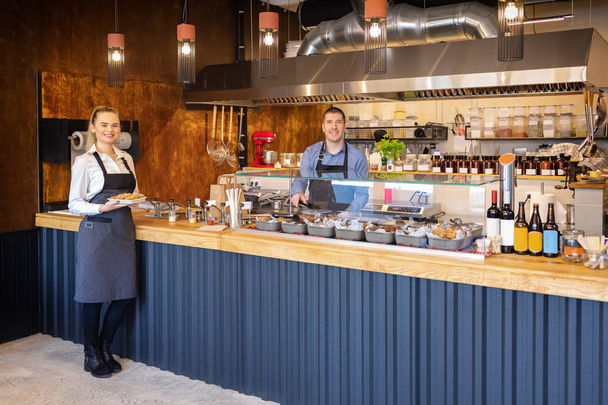 Υπηρεσία υποστήριξης σε μοντέρνο μπιστρό με χαμογελαστές σερβιτόρους που σερβίρουν φαγητό, χαρούμενους ιδιοκτήτες επιχειρήσεων σε μικρό εστιατόριο με ανοιχτή κουζίνα που προσκαλεί ανθρώπους μέσα για γευσιγνωσία φαγητού, υπηρεσία catering - Φωτογραφία, εικόνα