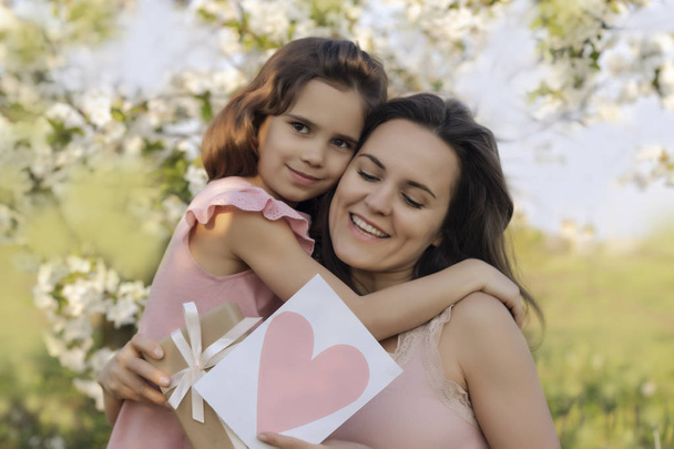 ¡Feliz Día de la Madre! hija le da a mamá una tarjeta de corazón y un regalo. Feliz Cumpleaños. Personas, vacaciones y familia concepto-feliz hija y madre en el contexto de flores en flor
 - Foto, imagen
