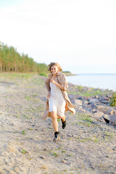 Young pretty woman walking on shingle beach and wearing summer coat. - Photo, Image