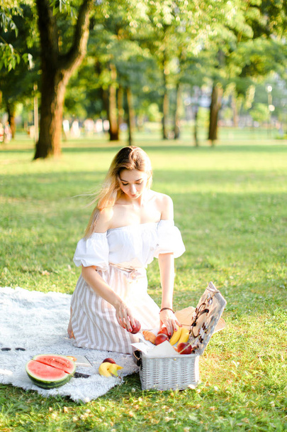 Молодая блондинка готовится к пикнику в парке, сидит на клетке возле коробки и ватерлимон
. - Фото, изображение