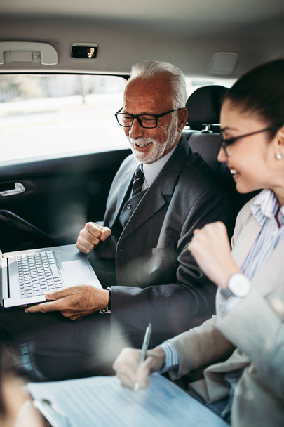 Good looking senior business man and his young woman colleague or coworker sitting on backseat in luxury car. They talking, smiling and using laptop and smart phones. Transportation in corporate business concept. - 写真・画像